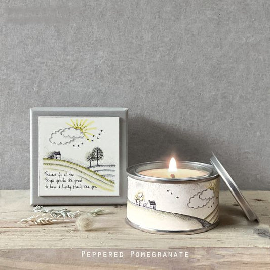 Candle in a tin with a decorative label on a wooden surface, against a gray wall.
