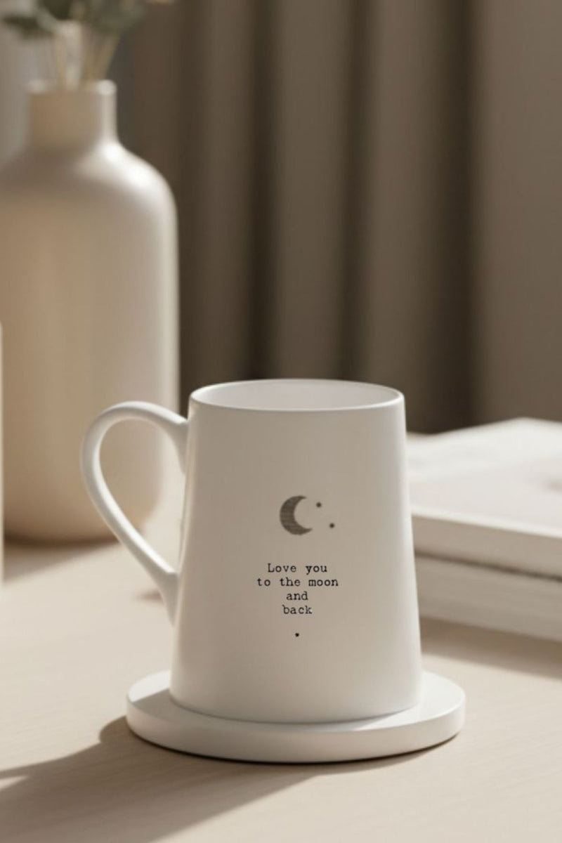 White ceramic mug with crescent moon and Love you to the moon and back text beside cream pillar candles, minimalist neutral decor.