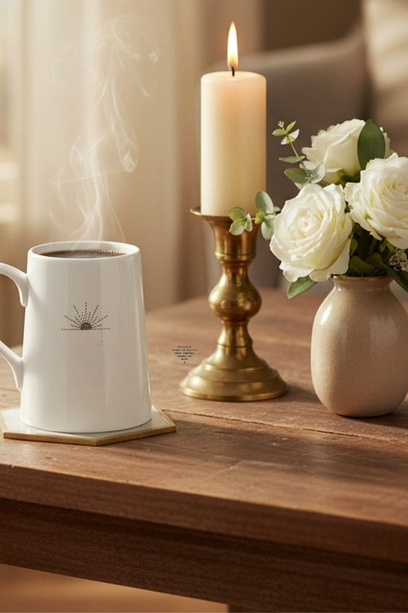 White quote mug “Distance means so little” on rustic wooden table beside lit candle and white rose vase in soft window light