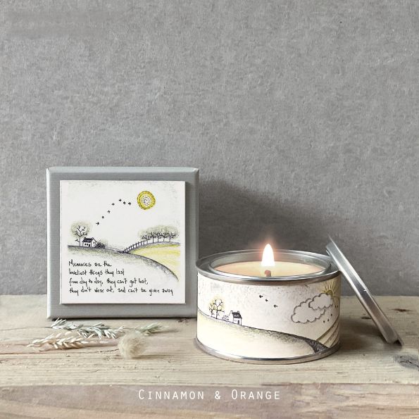Candle in a tin with a decorative label on a wooden surface, featuring a small framed artwork.