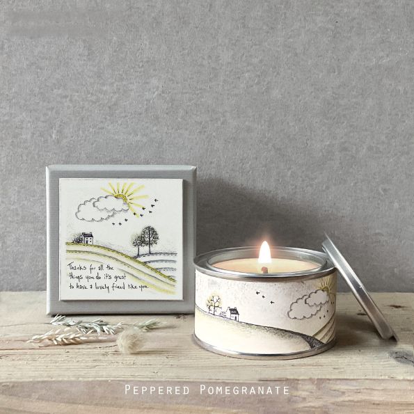Candle in a tin with a decorative label on a wooden surface, against a gray wall.