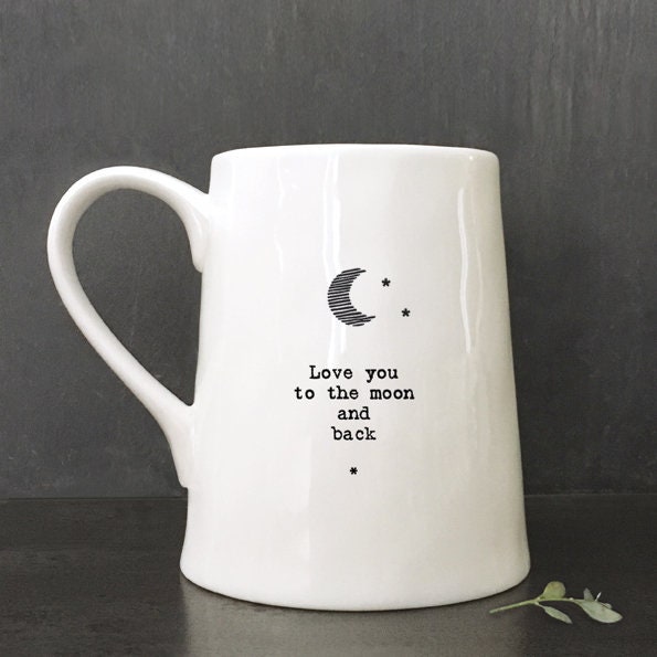White ceramic mug with minimalist black text and a crescent moon illustration reading “Love you to the moon and back,” photographed against a dark slate background with a small green leaf beside the base.
