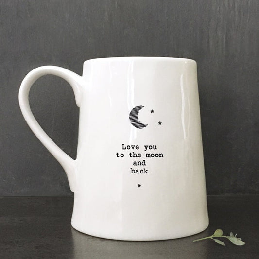 White ceramic mug with minimalist black text and a crescent moon illustration reading “Love you to the moon and back,” photographed against a dark slate background with a small green leaf beside the base.