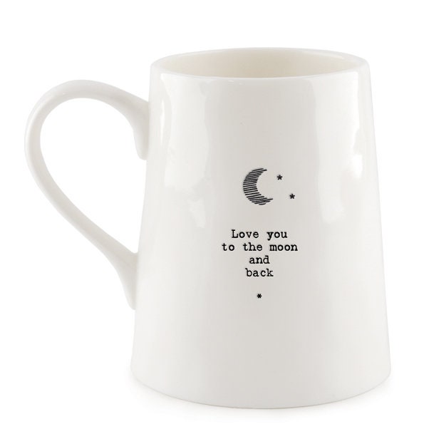White ceramic mug with minimalist black text and a crescent moon illustration reading “Love you to the moon and back,” photographed against a dark slate background with a small green leaf beside the base