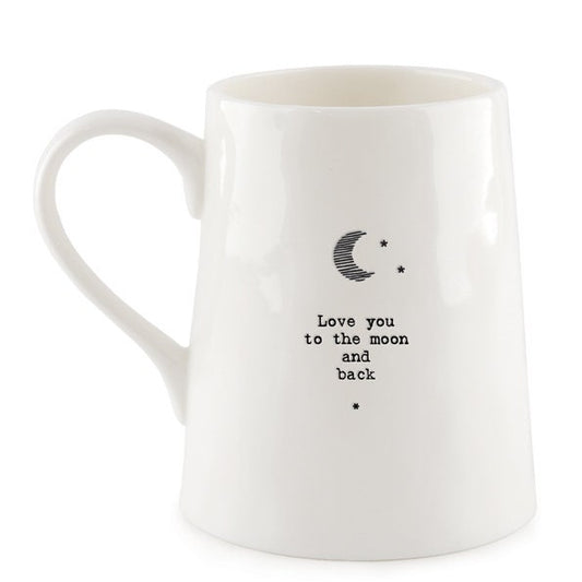 White ceramic mug with minimalist black text and a crescent moon illustration reading “Love you to the moon and back,” photographed against a dark slate background with a small green leaf beside the base