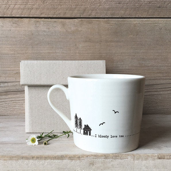 White mug with decorative text and design on a wooden surface with a box and flowers.