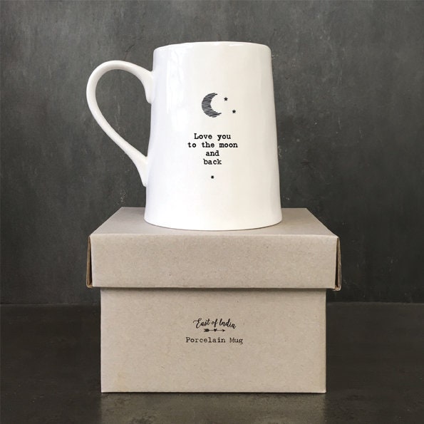 White ceramic mug with minimalist black text and a crescent moon illustration reading “Love you to the moon and back,” photographed against a dark slate background with a small green leaf beside the base