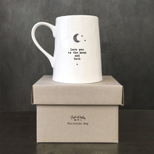 White ceramic mug with minimalist black text and a crescent moon illustration reading “Love you to the moon and back,” photographed against a dark slate background with a small green leaf beside the base