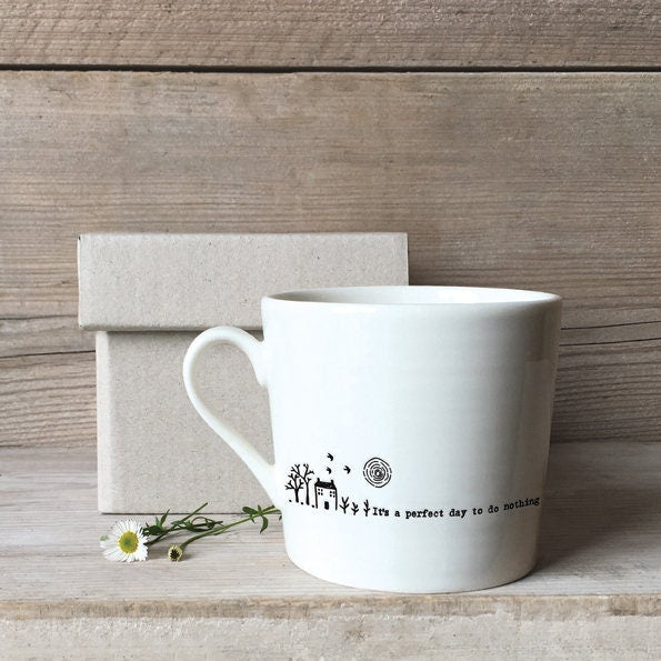 White mug with text and design on a wooden surface with a box and flower.