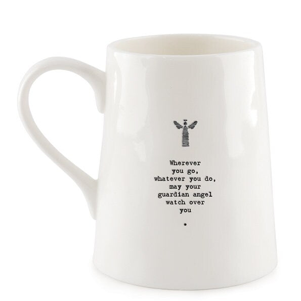 White ceramic mug with minimalist black text and a small angel illustration reading “Wherever you go, whatever you do, may your guardian angel watch over you,” photographed against a dark slate background with a small green leaf beside the base.