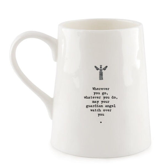 White ceramic mug with minimalist black text and a small angel illustration reading “Wherever you go, whatever you do, may your guardian angel watch over you,” photographed against a dark slate background with a small green leaf beside the base.