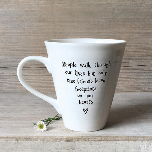 White ceramic friendship mug with black handwritten quote “People walk through our lives but only true friends leave footprints on our hearts,” displayed on a rustic wooden background with a small white daisy flower beside the base.